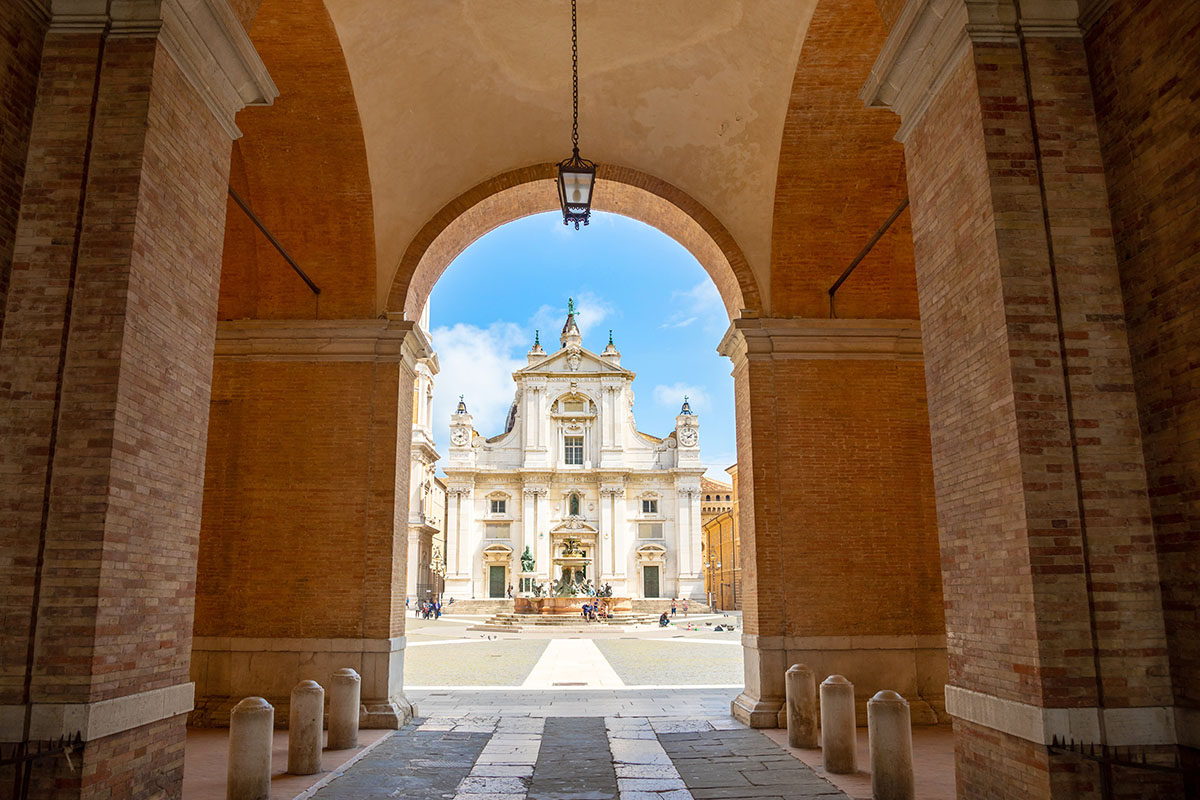 Loreto, Ancona, Italy – 8.05.2018: Square of Loreto with background the basilica in sunny day, portico to the side, people in the square in Loreto